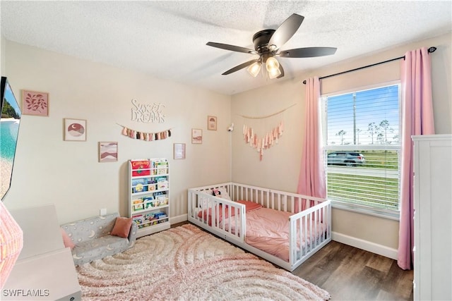 bedroom featuring a ceiling fan, wood finished floors, baseboards, a nursery area, and a textured ceiling