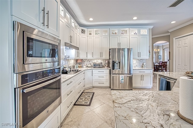 kitchen with appliances with stainless steel finishes, ornamental molding, light stone countertops, white cabinets, and decorative backsplash