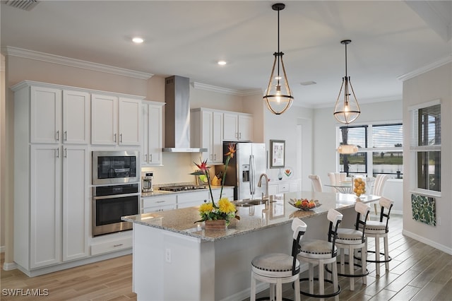 kitchen with pendant lighting, wall chimney range hood, appliances with stainless steel finishes, white cabinetry, and a kitchen island with sink