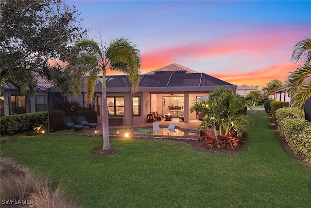 back house at dusk with a patio, glass enclosure, and a lawn