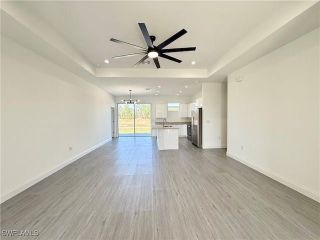 unfurnished living room with ceiling fan with notable chandelier, a tray ceiling, sink, and light hardwood / wood-style flooring