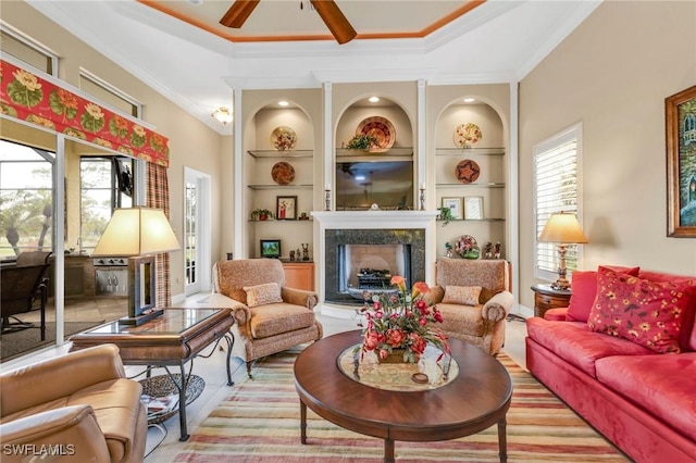 living room featuring built in features, a premium fireplace, ceiling fan, a tray ceiling, and ornamental molding