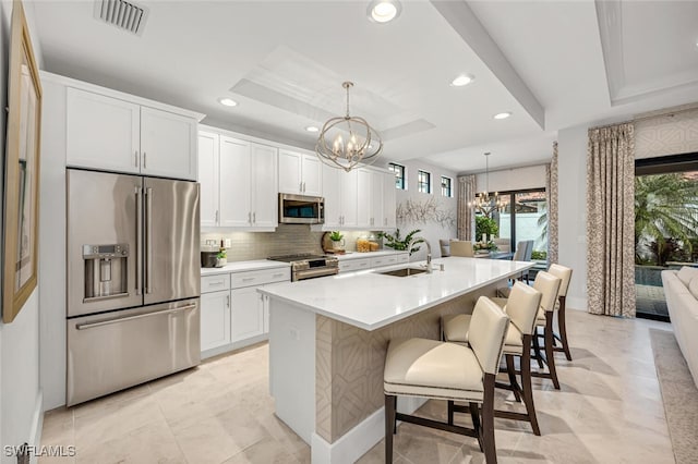 kitchen with appliances with stainless steel finishes, a notable chandelier, a tray ceiling, a center island with sink, and decorative light fixtures