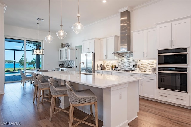 kitchen with wall chimney range hood, an island with sink, white cabinets, and appliances with stainless steel finishes