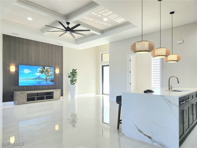 kitchen with sink, coffered ceiling, light stone counters, an island with sink, and decorative light fixtures