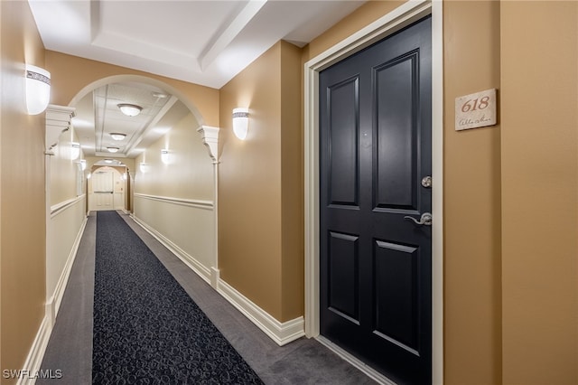 corridor featuring a tray ceiling, decorative columns, and dark colored carpet