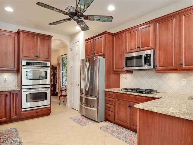 kitchen with light tile patterned floors, ceiling fan, appliances with stainless steel finishes, backsplash, and light stone counters