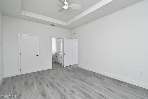 unfurnished bedroom with ceiling fan, ensuite bathroom, a tray ceiling, and light wood-type flooring