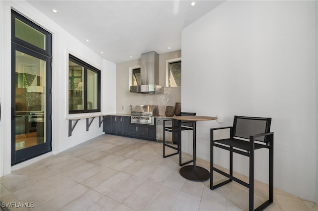 interior space featuring tasteful backsplash, wall chimney range hood, light tile patterned floors, and a breakfast bar area