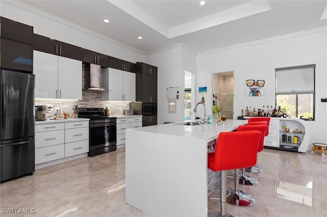 kitchen featuring an island with sink, a breakfast bar area, black appliances, a raised ceiling, and wall chimney range hood