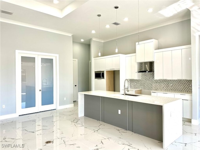 kitchen featuring wall chimney range hood, sink, white cabinetry, hanging light fixtures, and a kitchen island with sink