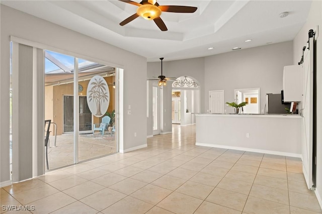 tiled empty room with a tray ceiling, a barn door, ceiling fan, and a towering ceiling