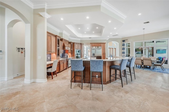 kitchen with a breakfast bar, built in appliances, a large island with sink, ornamental molding, and a raised ceiling