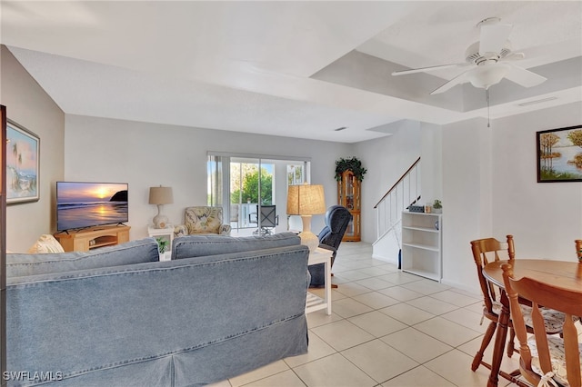 living area with a tray ceiling, stairway, light tile patterned floors, and a ceiling fan