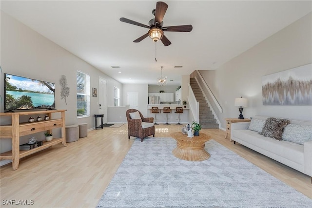 living area featuring light wood-style floors, stairway, baseboards, and ceiling fan with notable chandelier