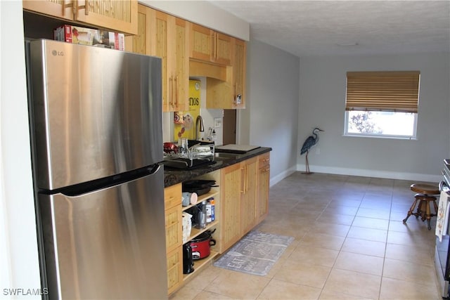 kitchen featuring light brown cabinetry, stainless steel refrigerator, sink, dark stone countertops, and light tile patterned floors