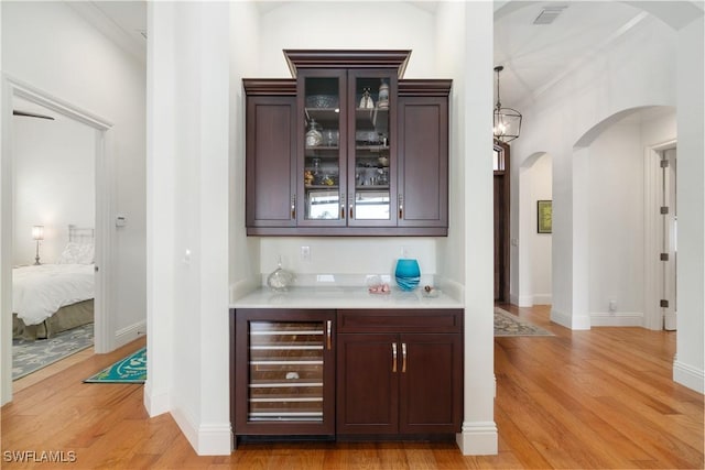 bar featuring wine cooler, dark brown cabinetry, hanging light fixtures, and light hardwood / wood-style flooring