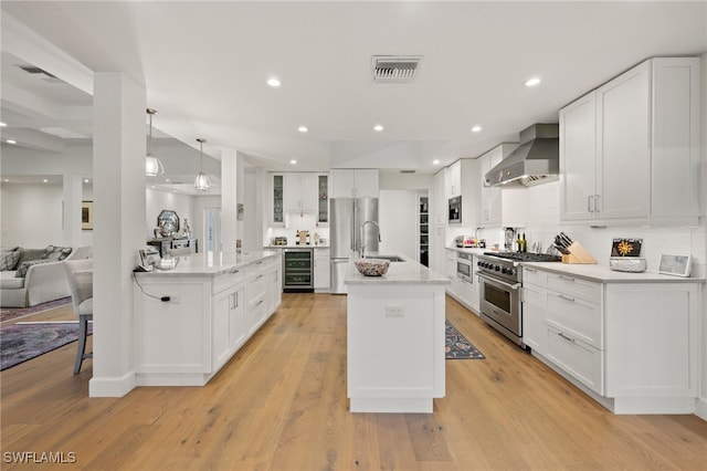 kitchen featuring a center island with sink, hanging light fixtures, stainless steel appliances, white cabinets, and wall chimney exhaust hood