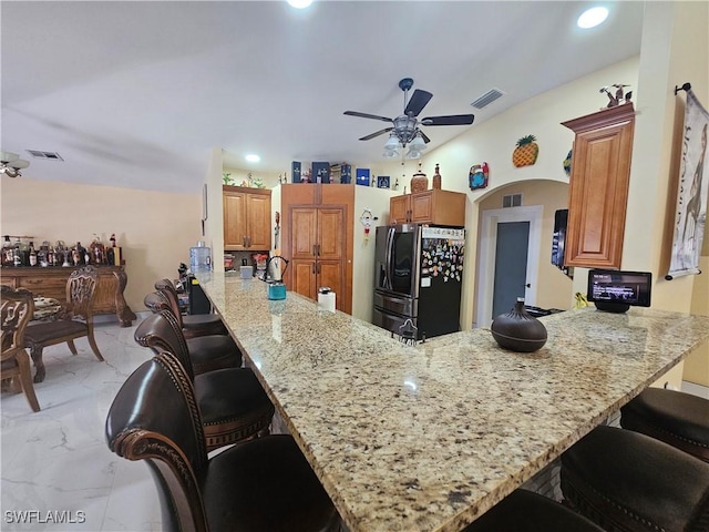 kitchen with kitchen peninsula, a kitchen breakfast bar, ceiling fan, light stone counters, and black fridge with ice dispenser