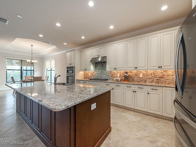 kitchen with decorative light fixtures, a tray ceiling, an island with sink, stainless steel appliances, and white cabinets