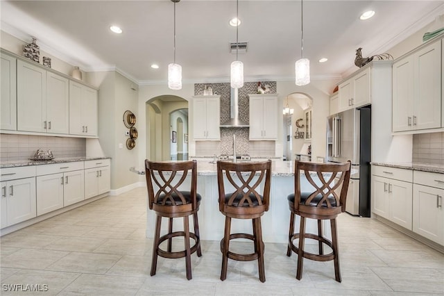 kitchen featuring hanging light fixtures, high quality fridge, an island with sink, light stone countertops, and wall chimney exhaust hood