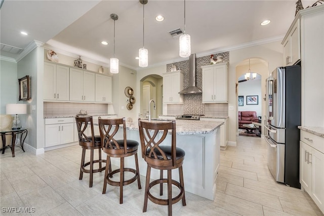 kitchen with stainless steel appliances, light stone countertops, a center island with sink, decorative light fixtures, and wall chimney exhaust hood