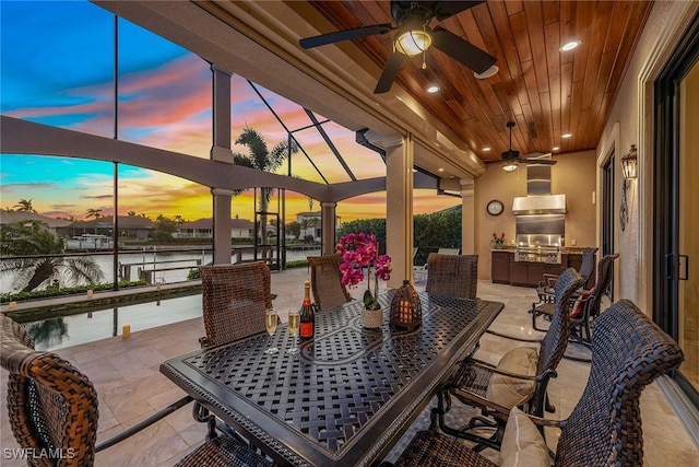 patio terrace at dusk featuring a water view, grilling area, exterior kitchen, and ceiling fan