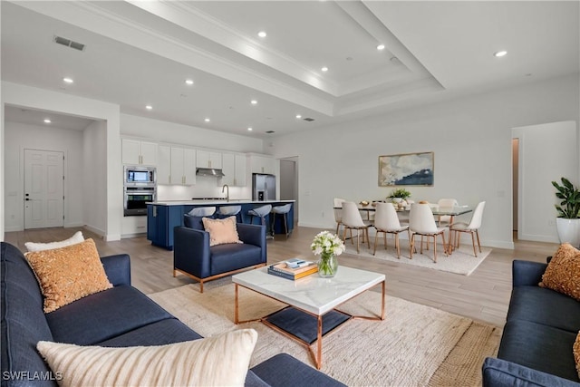 living room with sink, ornamental molding, a raised ceiling, and light wood-type flooring