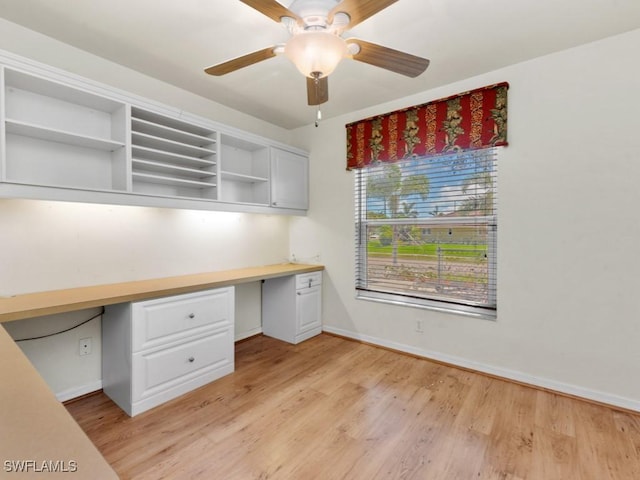 unfurnished office featuring light wood-style flooring, ceiling fan, built in desk, and baseboards