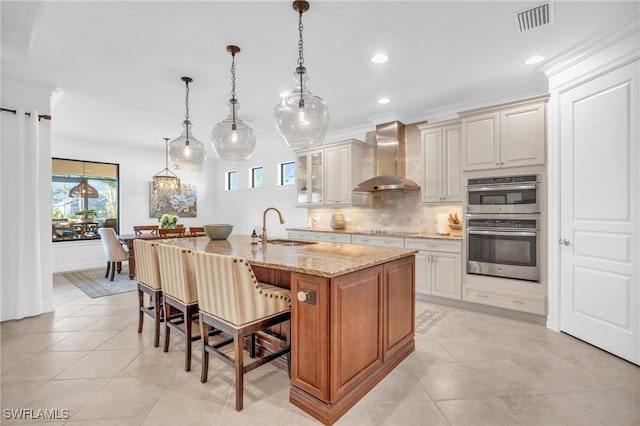 kitchen featuring pendant lighting, sink, a kitchen island with sink, light stone counters, and wall chimney range hood