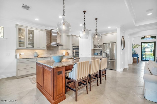 kitchen with pendant lighting, a kitchen island with sink, light stone counters, stainless steel fridge with ice dispenser, and wall chimney exhaust hood