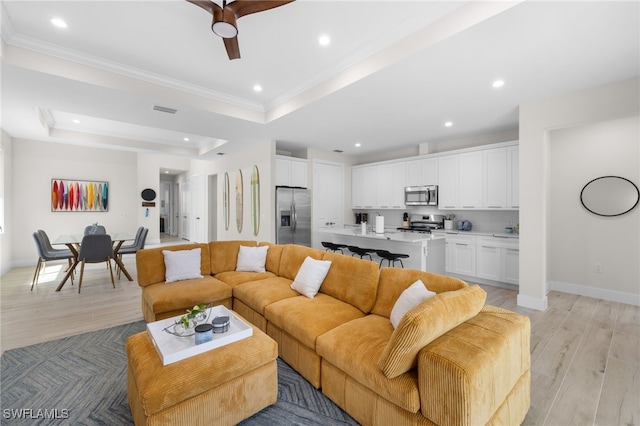 living room with ceiling fan, ornamental molding, a raised ceiling, and light wood-type flooring