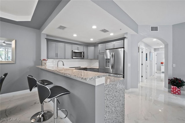 kitchen with appliances with stainless steel finishes, a breakfast bar area, gray cabinetry, light stone counters, and kitchen peninsula