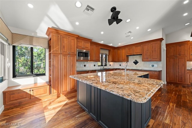 kitchen with stainless steel microwave, dark wood-type flooring, light stone countertops, and a large island with sink