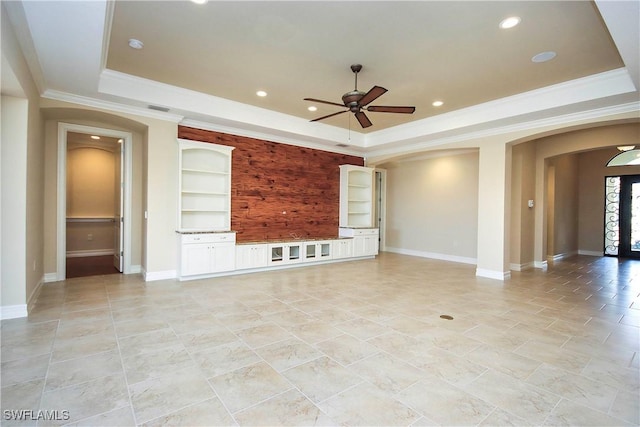 unfurnished living room with arched walkways, baseboards, built in features, a tray ceiling, and crown molding