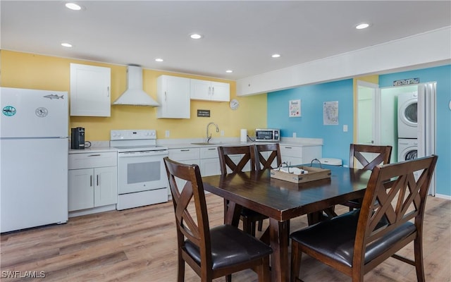 kitchen featuring wall chimney exhaust hood, stacked washer and clothes dryer, white cabinetry, light hardwood / wood-style flooring, and white appliances