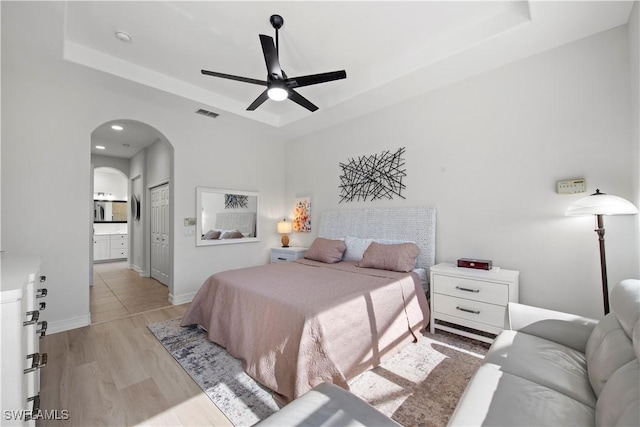 bedroom featuring light hardwood / wood-style flooring, ceiling fan, a tray ceiling, ensuite bath, and a closet