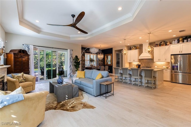 living area with visible vents, light wood-style flooring, ornamental molding, a tray ceiling, and ceiling fan with notable chandelier