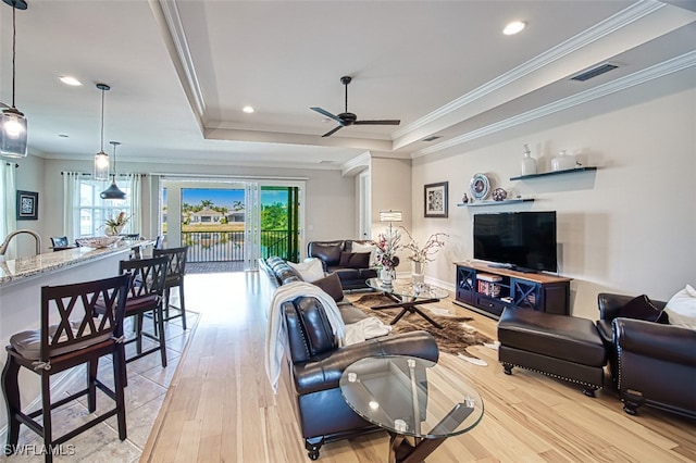 living area featuring visible vents, a ceiling fan, ornamental molding, a tray ceiling, and light wood-type flooring