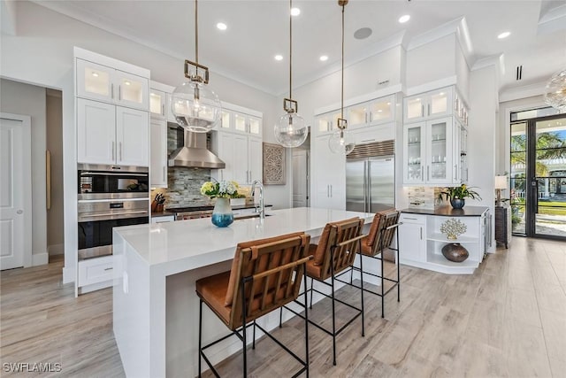 kitchen with white cabinetry, decorative light fixtures, wall chimney exhaust hood, and a center island with sink