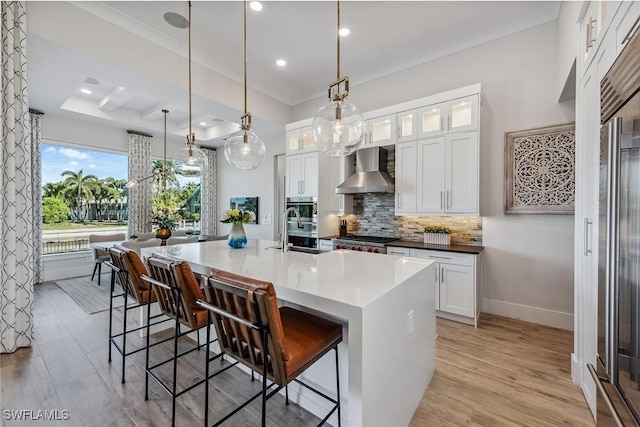 kitchen with a breakfast bar area, decorative light fixtures, wall chimney range hood, a kitchen island with sink, and white cabinets