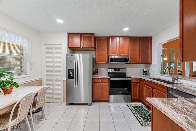 kitchen with light tile patterned floors, light stone countertops, stainless steel appliances, a sink, and brown cabinets