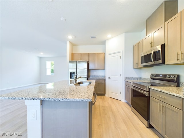 kitchen with appliances with stainless steel finishes, an island with sink, sink, light stone countertops, and light wood-type flooring