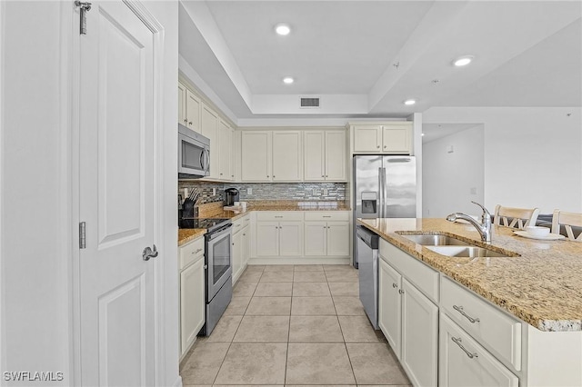 kitchen featuring appliances with stainless steel finishes, an island with sink, sink, light stone counters, and a tray ceiling