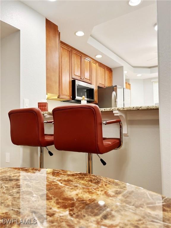 kitchen with light stone counters, a tray ceiling, a breakfast bar area, stainless steel appliances, and brown cabinetry