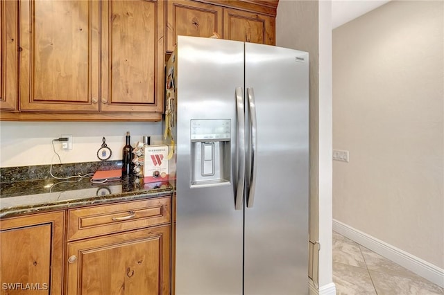 kitchen featuring light tile patterned floors, baseboards, stainless steel fridge with ice dispenser, dark stone counters, and brown cabinetry