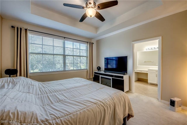 bedroom with ceiling fan, light colored carpet, a tray ceiling, and ensuite bath