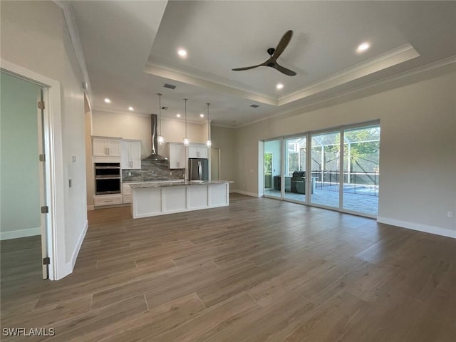 kitchen with wall chimney exhaust hood, appliances with stainless steel finishes, a tray ceiling, an island with sink, and white cabinets