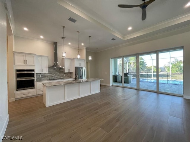 kitchen featuring wall chimney range hood, pendant lighting, stainless steel appliances, a kitchen island with sink, and white cabinets