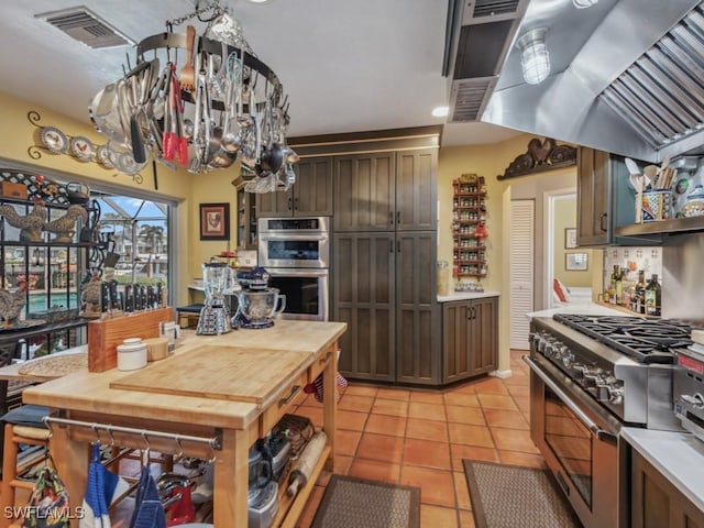 kitchen with dark brown cabinets, light tile patterned flooring, wall chimney exhaust hood, and appliances with stainless steel finishes